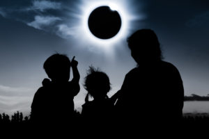 Natural phenomenon. Silhouette back view of mother and child sitting and relaxing together. Boy pointing to solar eclipse on dark sky background. Happy family spending time together. Outdoor.